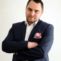 Portret mężczyzny w granatowym garniturze, białej koszuli i wzorzystej poszetce, z założonymi rękami na jasnym tle.