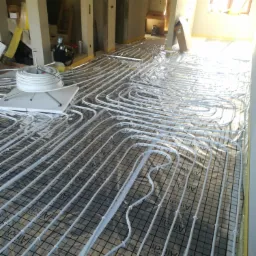 Rozłożone rury ogrzewania podłogowego na izolacji z siatką w surowym wnętrzu budynku w trakcie budowy, widoczny zwój rur i narzędzia w tle.