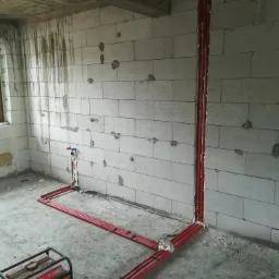 Czerwone rury instalacji wodnej prowadzone wzdłuż ściany z betonu komórkowego, z generatorem prądu Kraft&Dele ST1300 na podłodze, w surowym stanie wykończenia wnętrza.