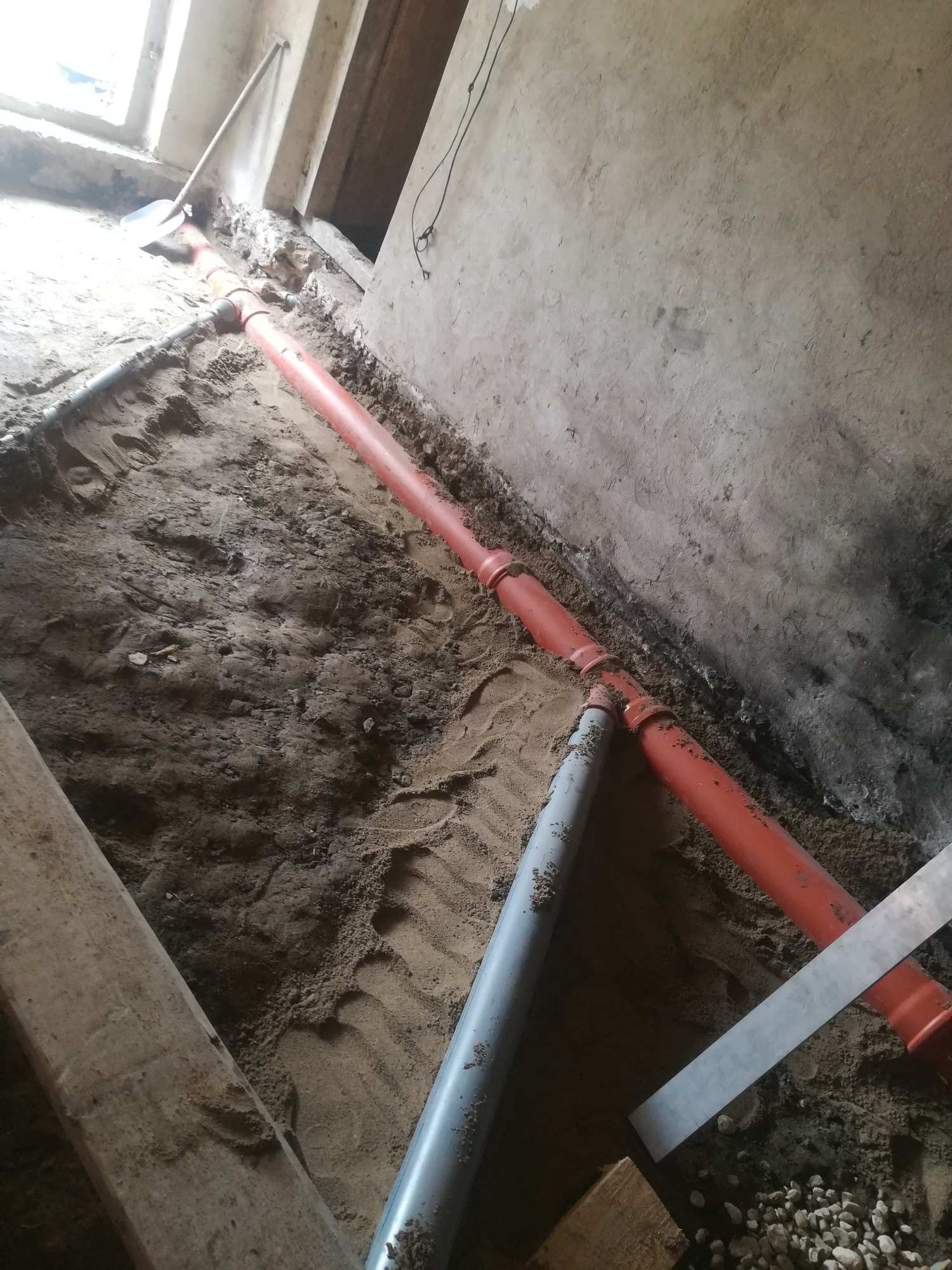 Wykop z ułożonymi rurami kanalizacyjnymi z tworzywa w kolorze szarym i pomarańczowym, widoczne złącza, piasek i gruz w wykopie, ściana z surowego betonu w tle.