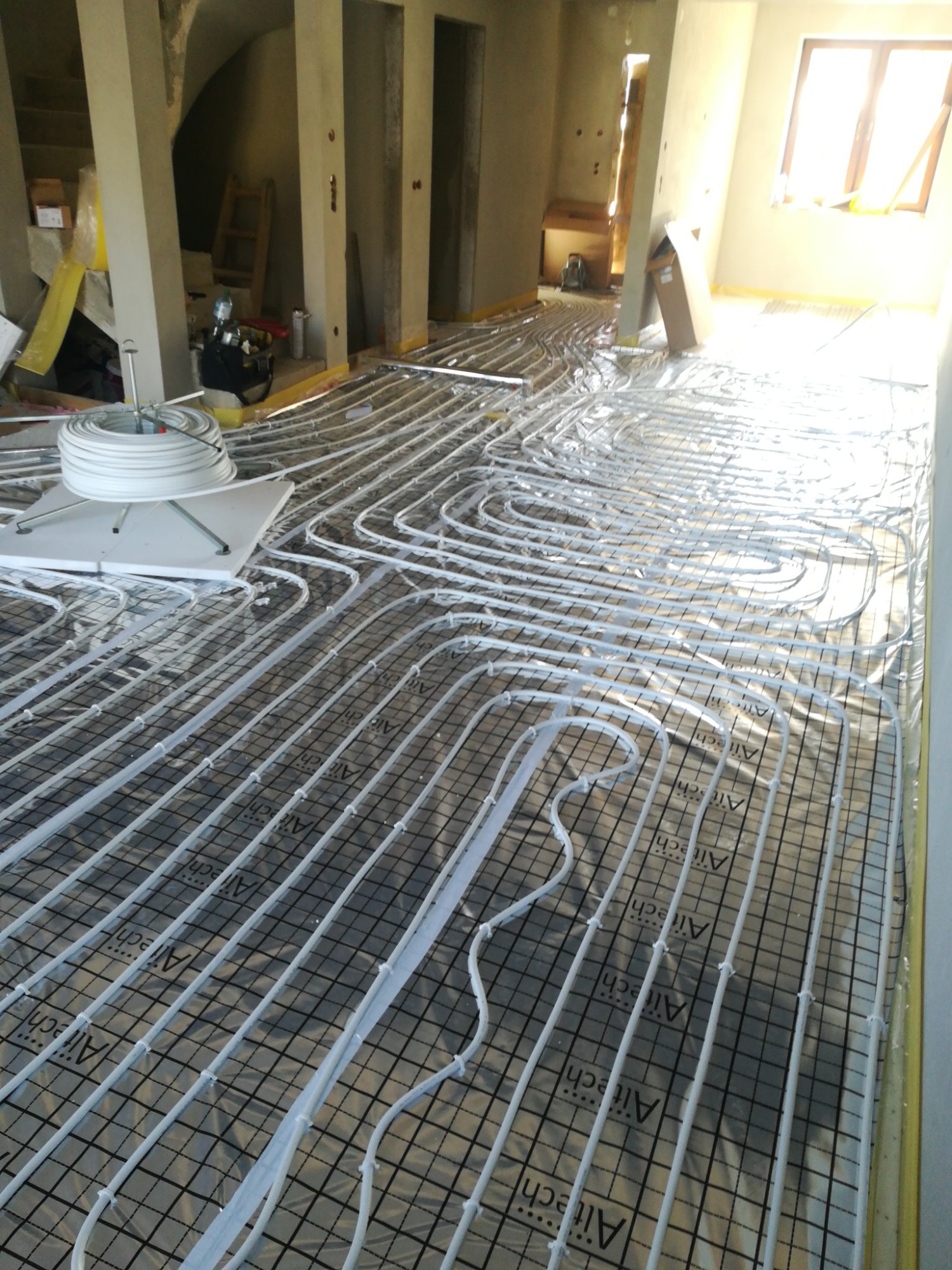 Rozłożone rury ogrzewania podłogowego na izolacji z siatką w surowym wnętrzu budynku w trakcie budowy, widoczny zwój rur i narzędzia w tle.