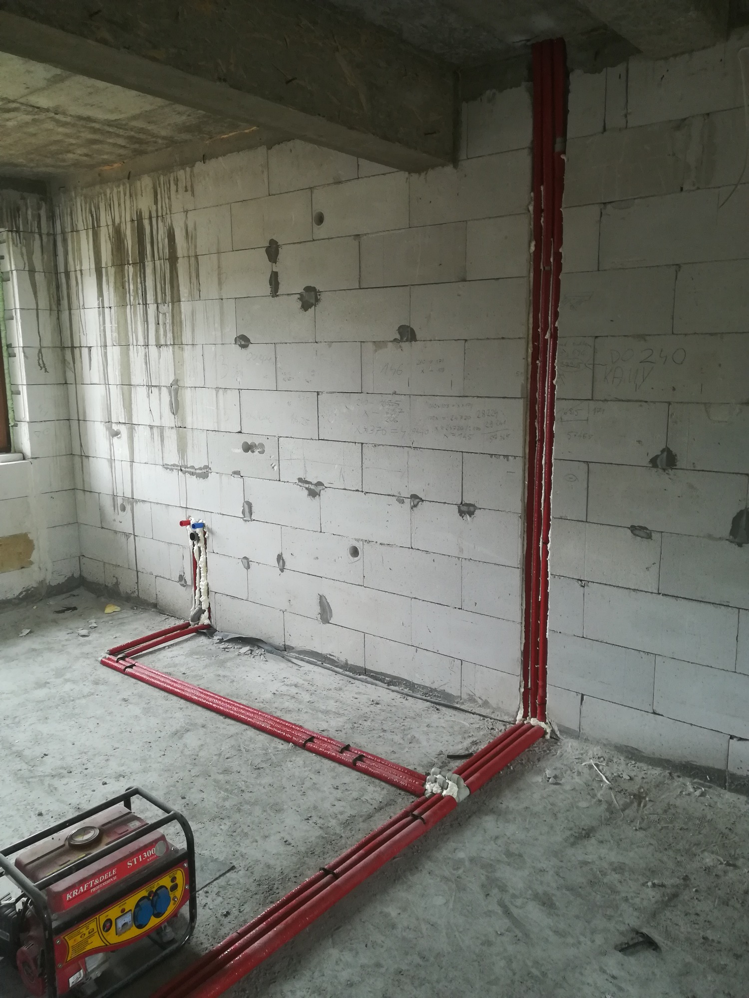 Czerwone rury instalacji wodnej prowadzone wzdłuż ściany z betonu komórkowego, z generatorem prądu Kraft&Dele ST1300 na podłodze, w surowym stanie wykończenia wnętrza.