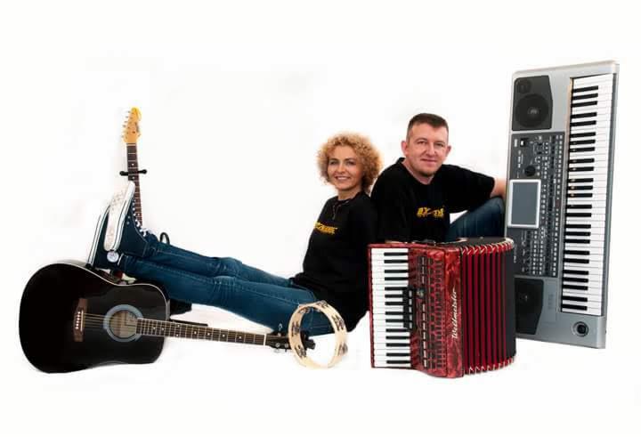 Dwoje muzyków pozuje z gitarą akustyczną, tamburynem, akordeonem i keyboardem na białym tle.