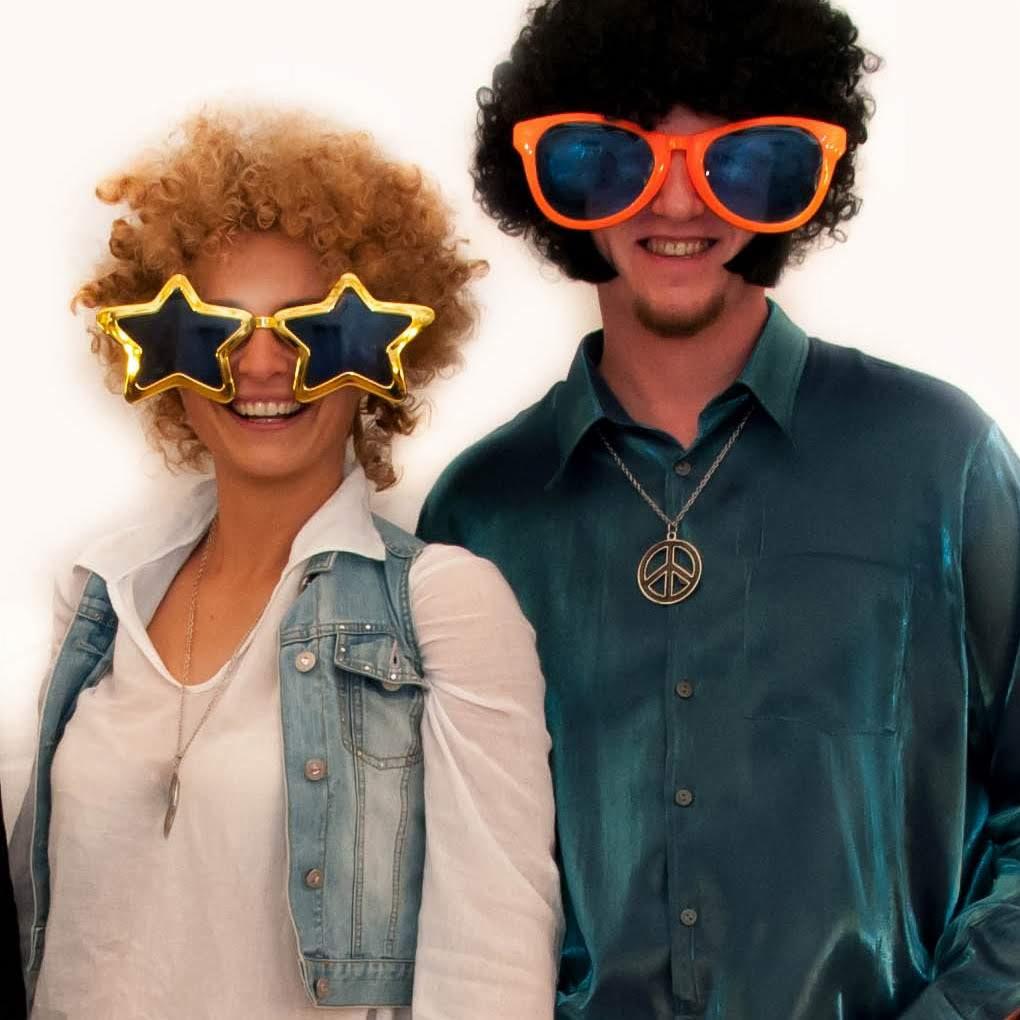 Para w stylizacjach disco: kobieta w blond afro, dżinsowej kamizelce i okularach w kształcie gwiazd, mężczyzna w czarnym afro, błyszczącej koszuli i pomarańczowych okularach, oboje z uśmiechem.