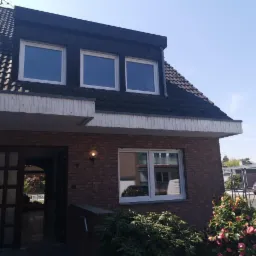 Dwukondygnacyjny dom z cegły z białymi oknami, widoczne trzy okna na poddaszu i jedno na parterze, otwarte drzwi frontowe z widokiem na wnętrze, krzewy z różowymi kwiatami w ogrodzie.