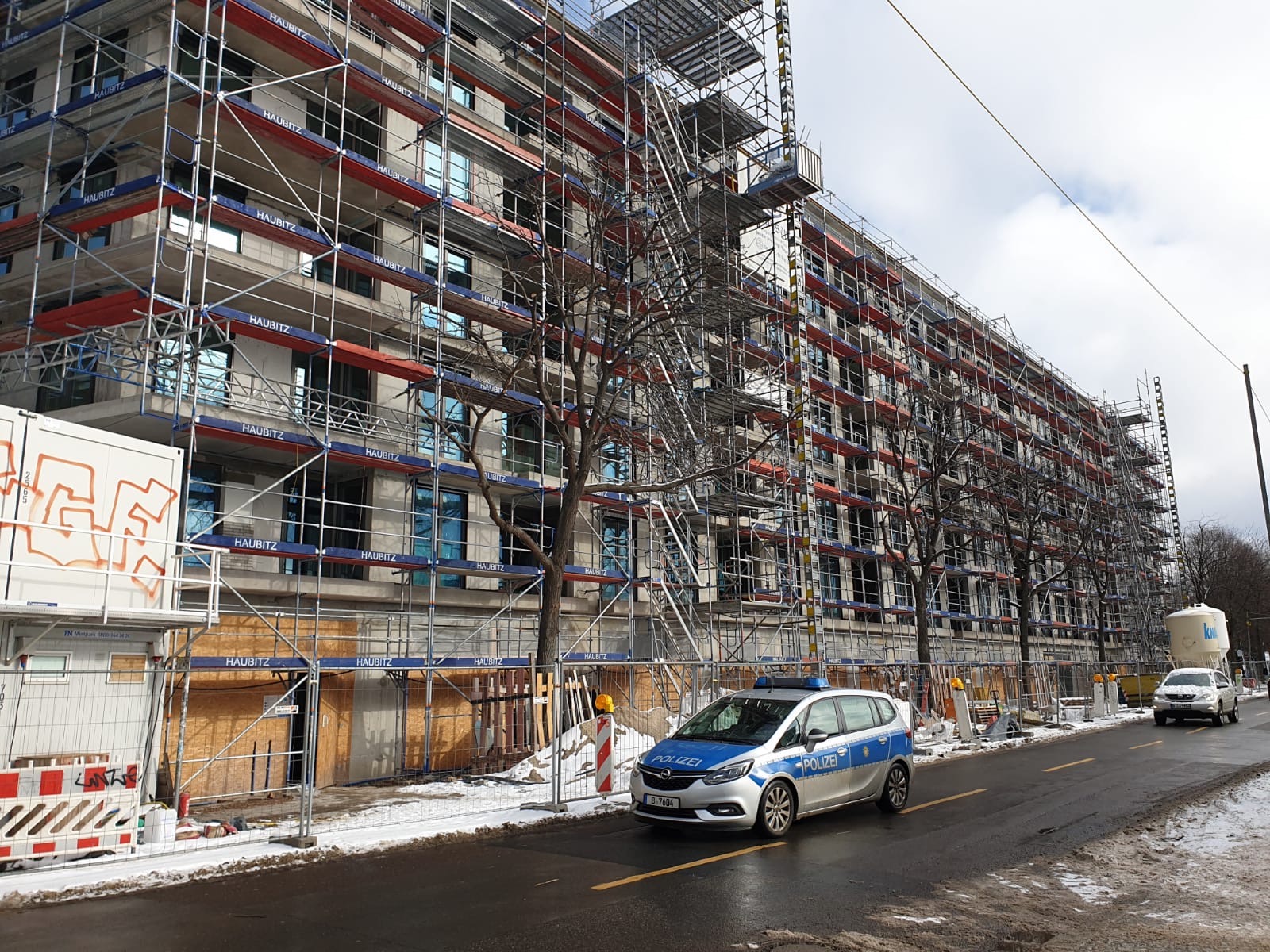 Budynek w trakcie remontu w Berlinie, otoczony rusztowaniami, z widocznymi nowymi, niebieskimi oknami. Na pierwszym planie zaparkowany samochód policyjny na mokrej ulicy.