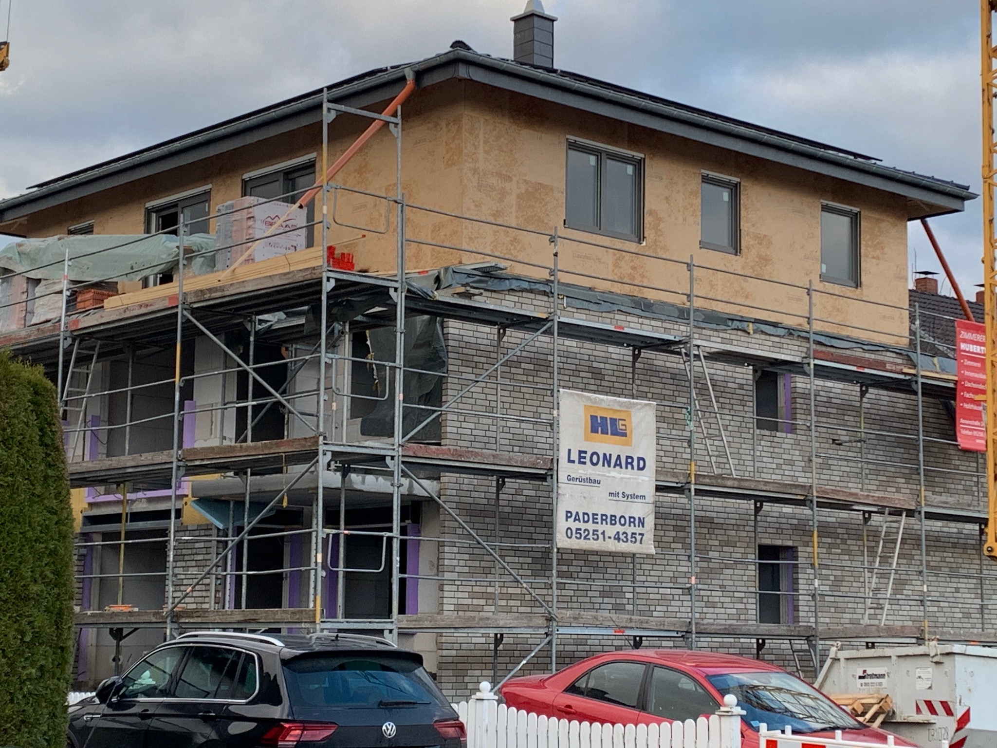 Budowa domu w Paderborn: rusztowania, cegły, okna, ocieplenie, samochody na pierwszym planie, widoczny baner firmy budowlanej.