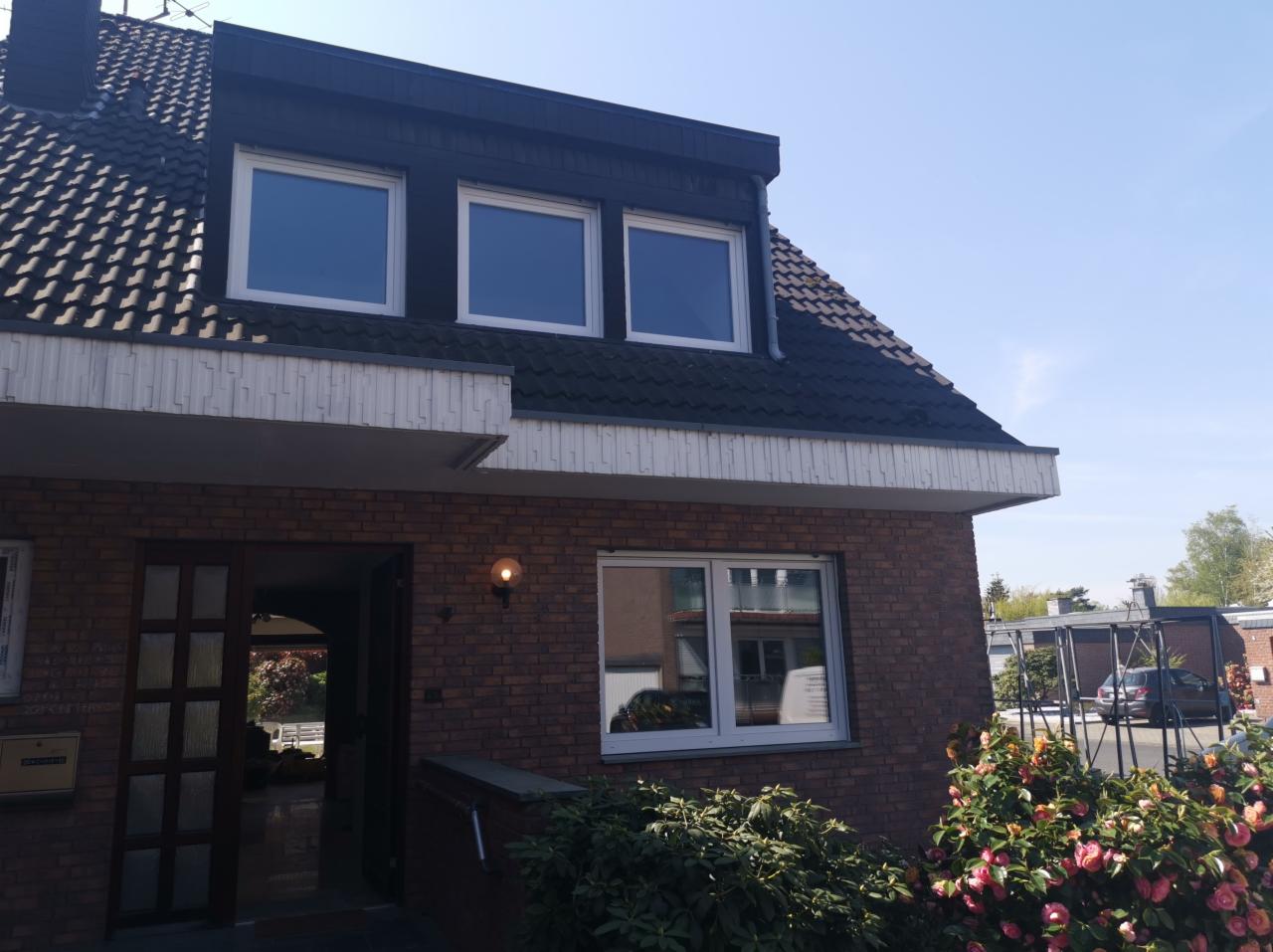 Dwukondygnacyjny dom z cegły z białymi oknami, widoczne trzy okna na poddaszu i jedno na parterze, otwarte drzwi frontowe z widokiem na wnętrze, krzewy z różowymi kwiatami w ogrodzie.