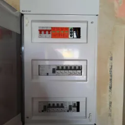 Nowa, biała skrzynka elektryczna z zamontowanymi bezpiecznikami i ogranicznikami przepięć, zamontowana na ścianie w trakcie remontu, widoczne ślady po starej farbie.