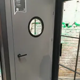 Szare drzwi z okrągłym okienkiem i czarną klamką, etykieta z informacjami o modelu drzwi, widok na ceglaną ścianę przez okno obok.