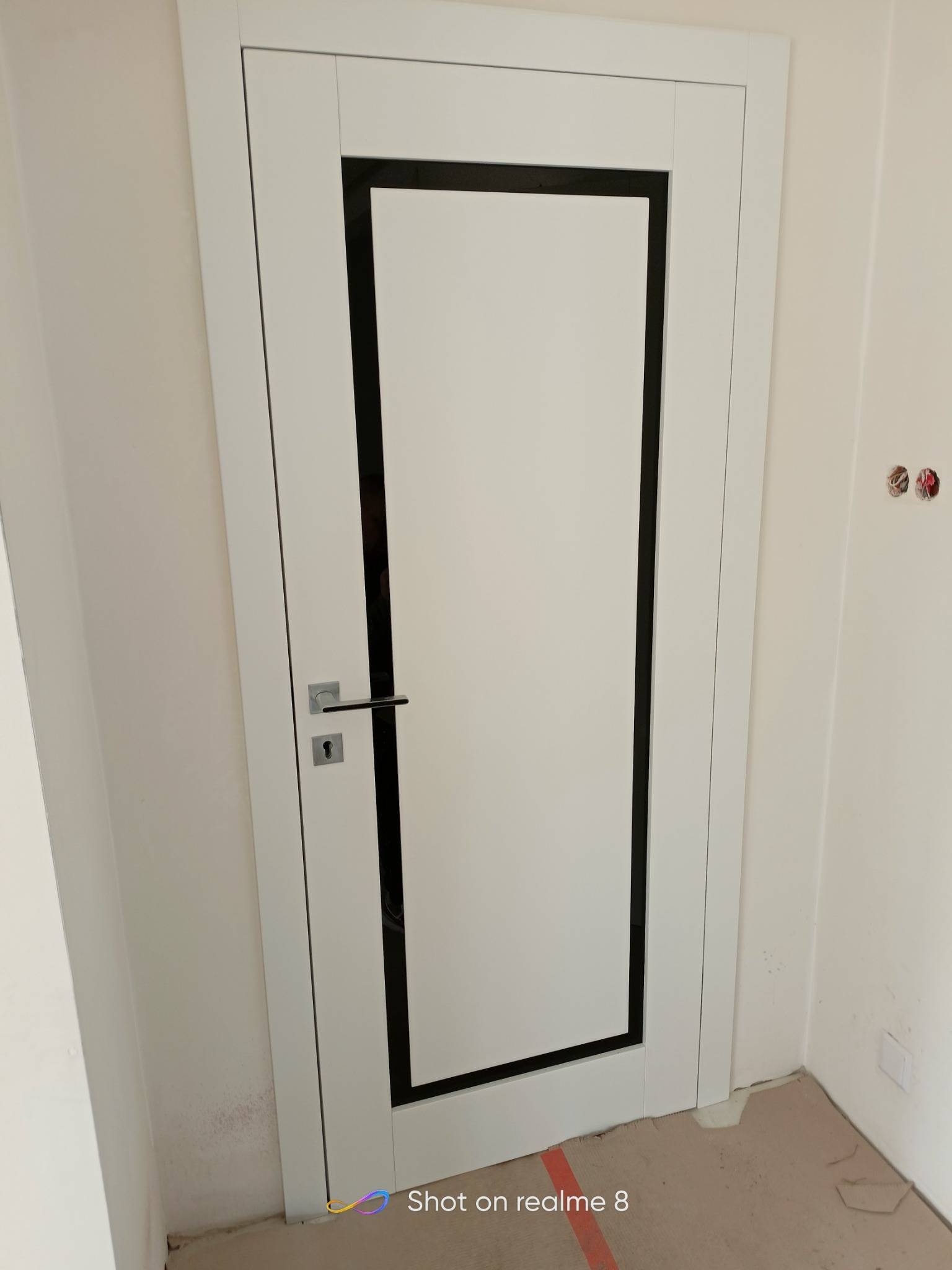 Białe drzwi wewnętrzne z czarną obwódką, zamontowane w surowym wnętrzu z widocznymi niedoskonałościami ścian i podłogi zabezpieczonej kartonem.