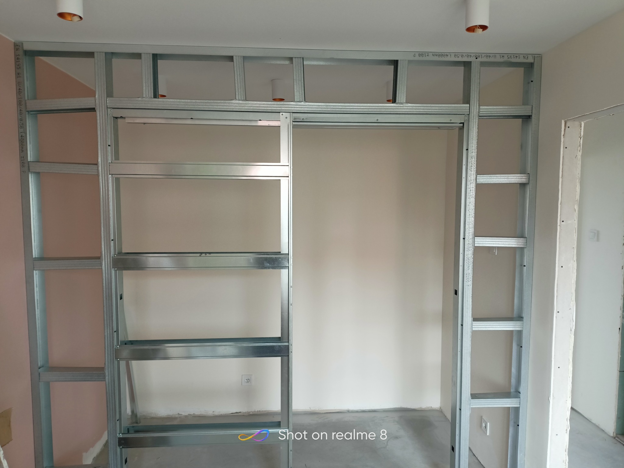 Metalowa konstrukcja szkieletowa ścianki działowej z profilem sufitowym i oświetleniem, widoczne kable elektryczne, ściana pomalowana na jasny kolor