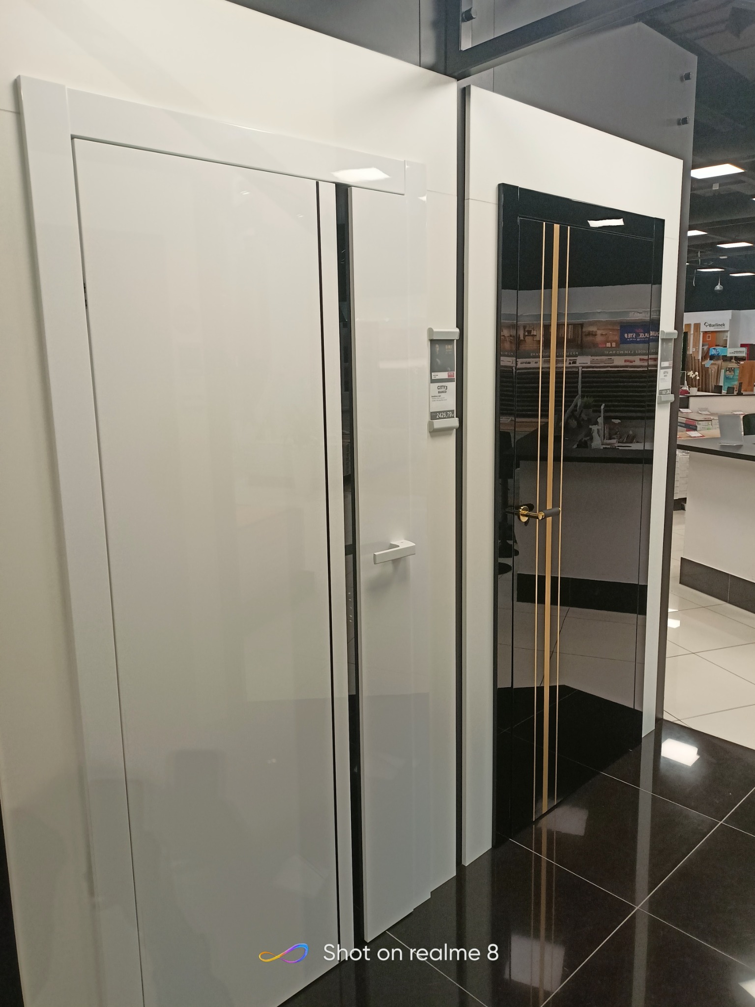 Ekspozycja dwóch modeli drzwi: białe skrzydło z minimalistyczną klamką oraz czarne, lakierowane drzwi z trzema złotymi paskami i elegancką klamką, prezentowane w salonie sprzedaży.