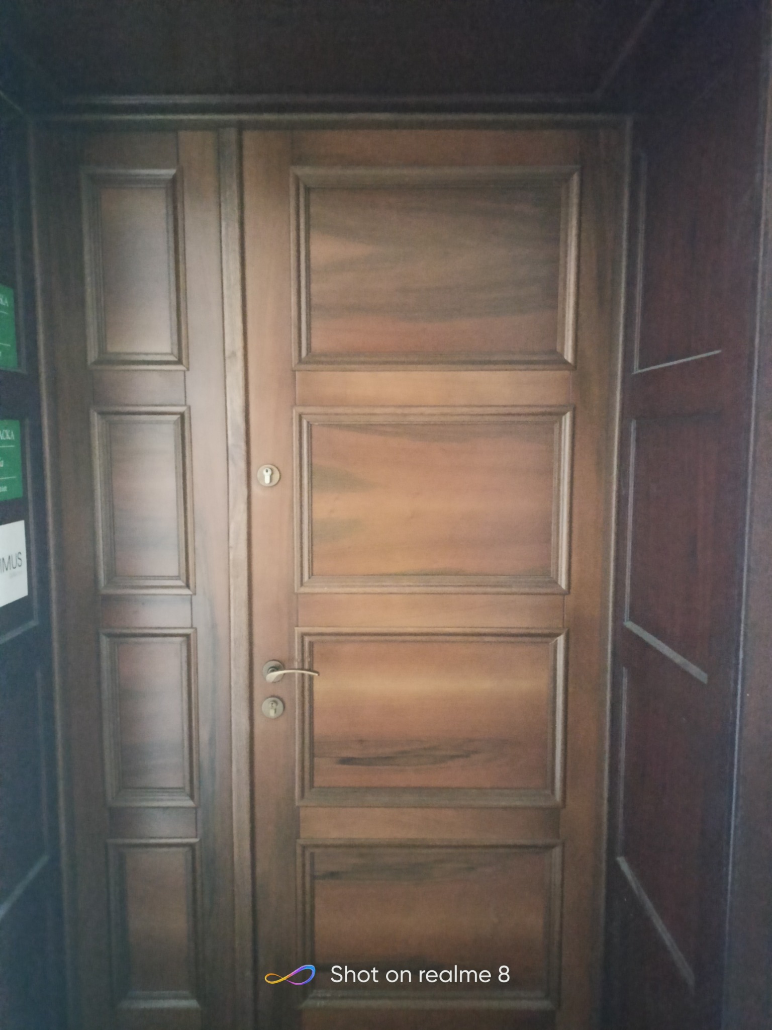 Dwuskrzydłowe drzwi wewnętrzne z drewna o ciepłym odcieniu, z widoczną klamką i zamkiem, wkomponowane w drewnianą boazerię.