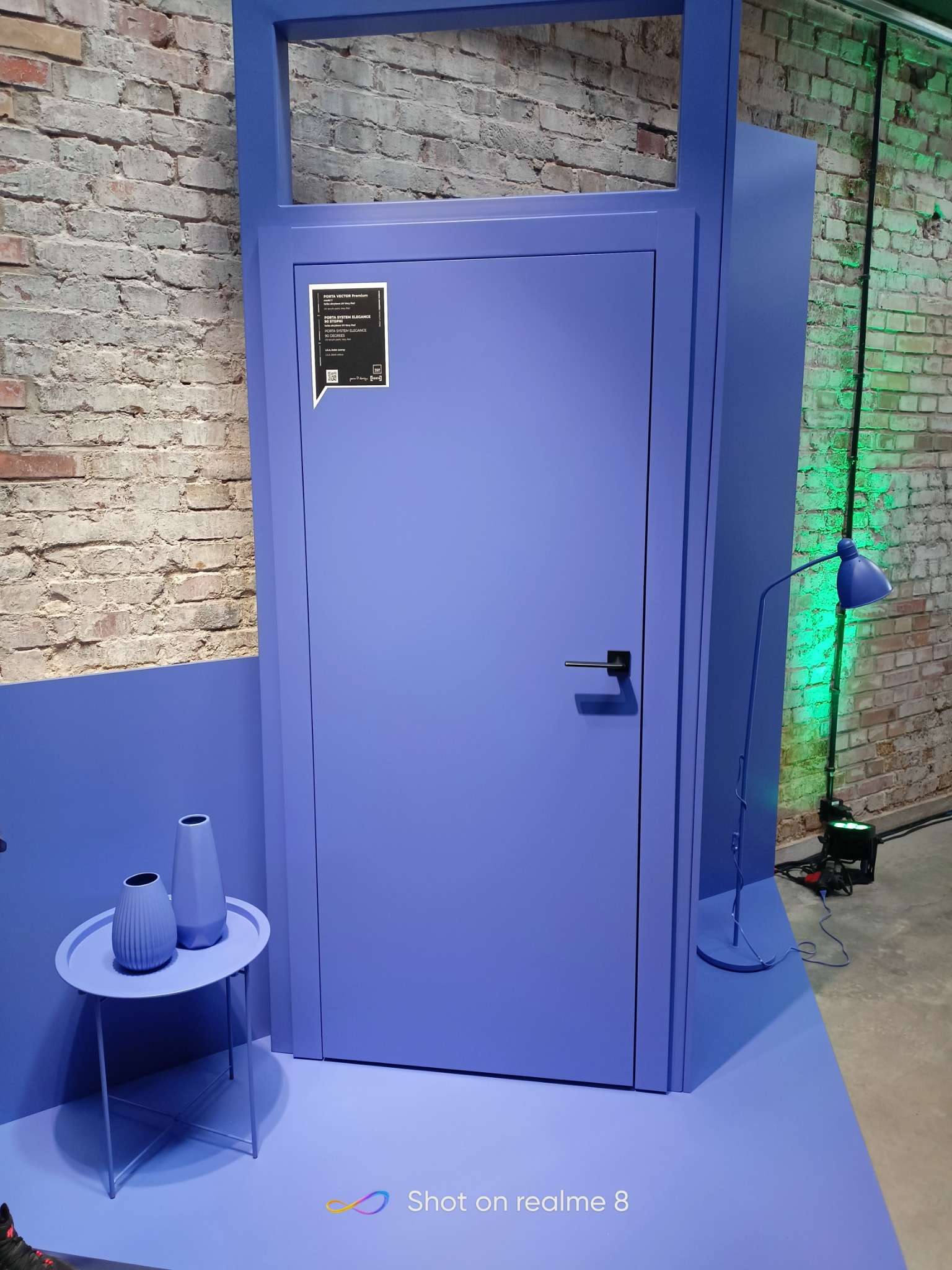 Niebieskie drzwi w minimalistycznej aranżacji, z lampą i stolikiem z wazonami, na tle ceglanej ściany oświetlonej zielonym światłem.