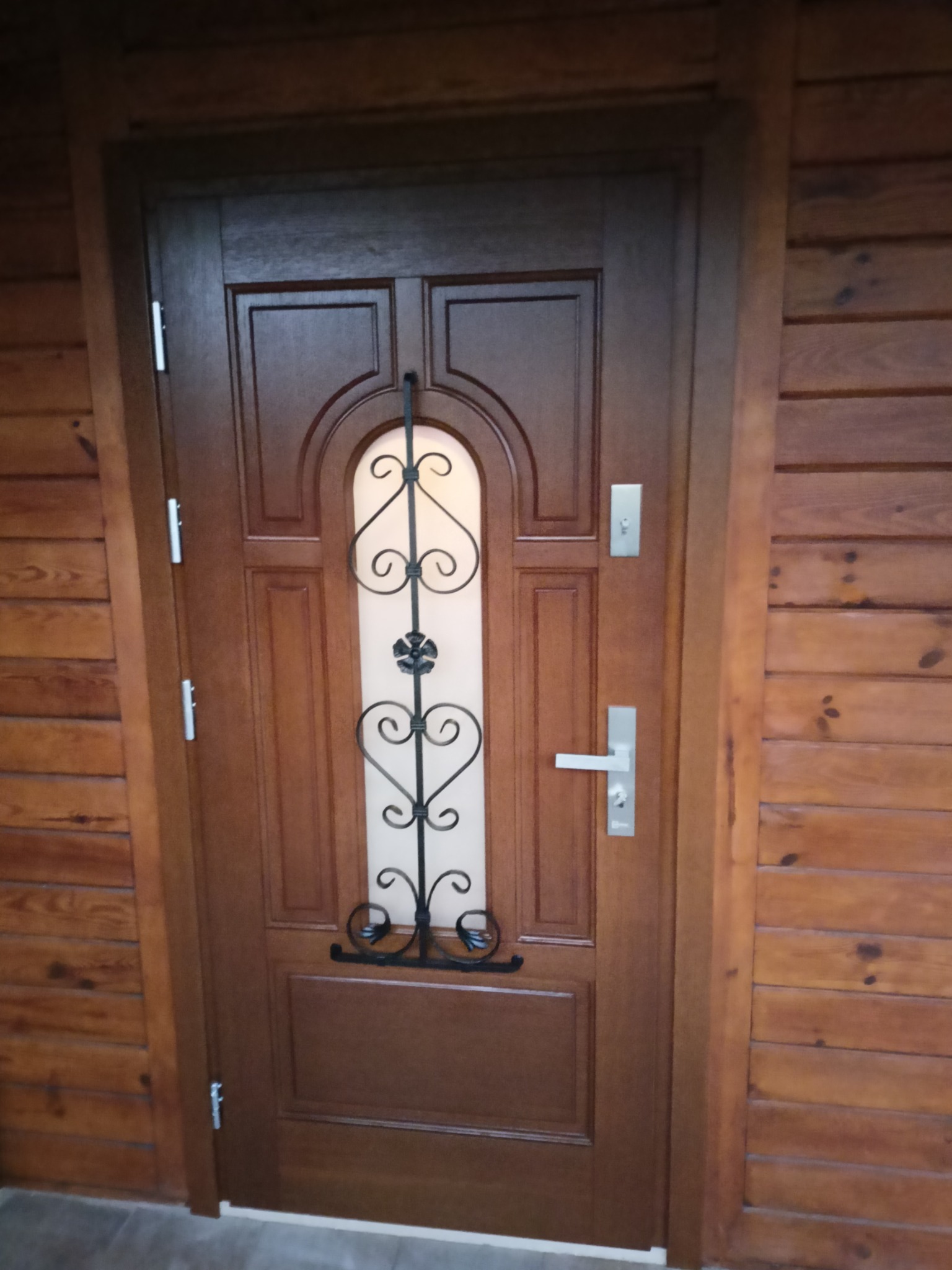 Brązowe drzwi wejściowe z dekoracyjną, kutą kratą w kształcie serc i kwiatu, zamontowane w drewnianej ścianie z boazerii.