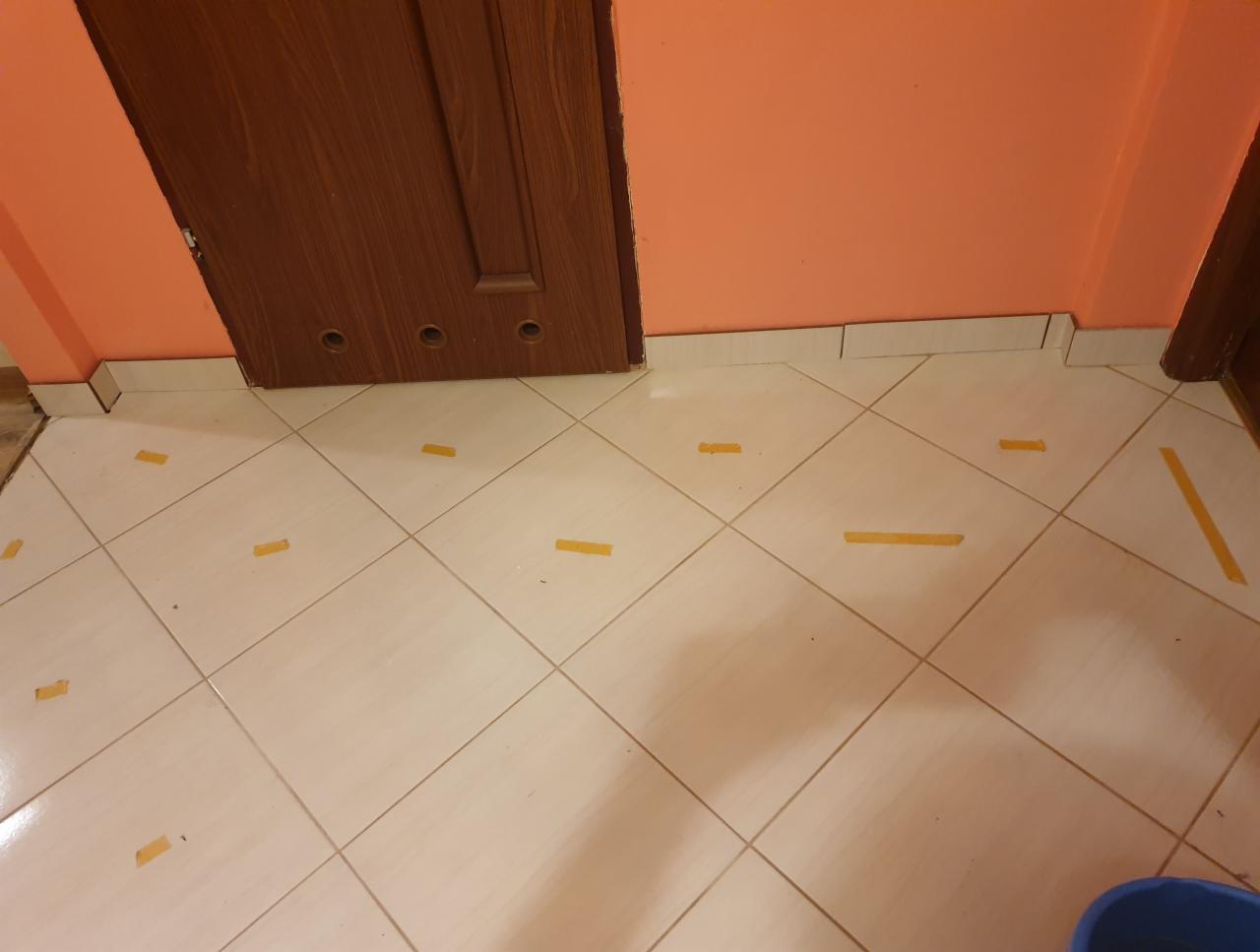 Podłoga wyłożona jasnymi, kwadratowymi płytkami, zabezpieczona żółtą taśmą malarską, widoczna pomarańczowa ściana i fragment brązowych drzwi.