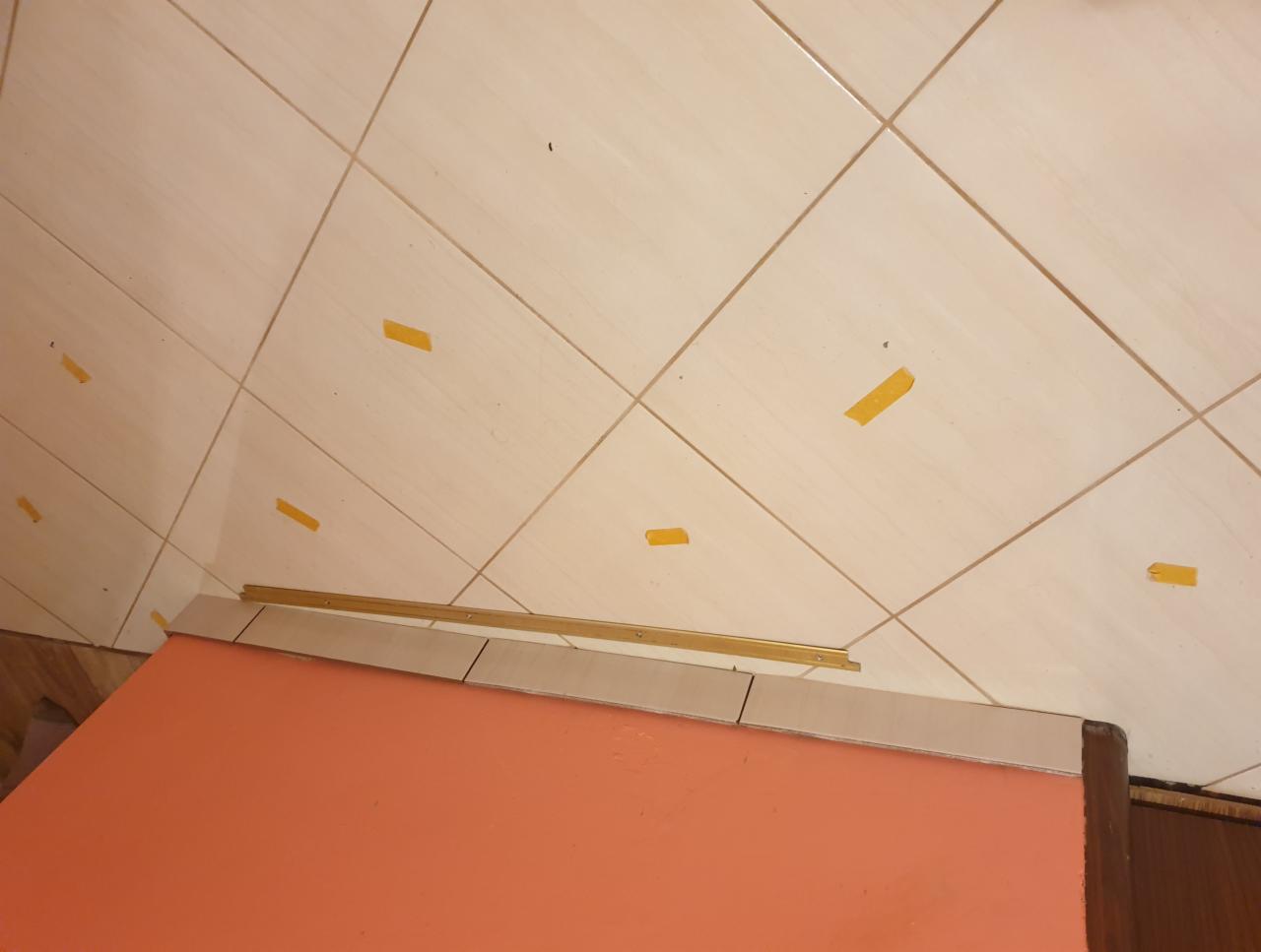 Beżowe płytki podłogowe ułożone ukośnie, tymczasowo zabezpieczone żółtą taśmą, widoczne listwy przypodłogowe i fragment pomarańczowej ściany.