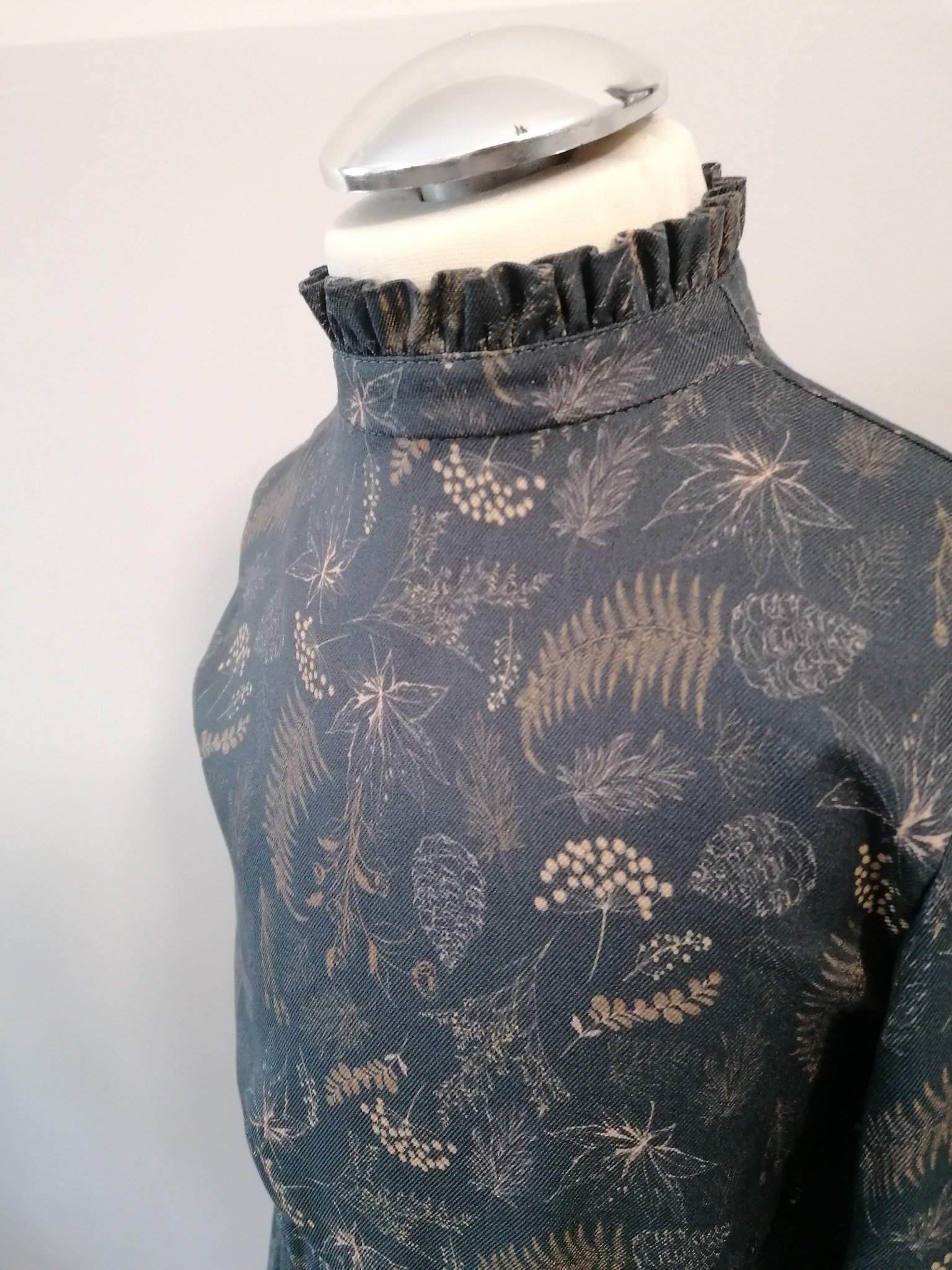 Bluzka z wysokim, marszczonym kołnierzem na manekinie krawieckim, wzór w liście i kwiaty na ciemnym tle.