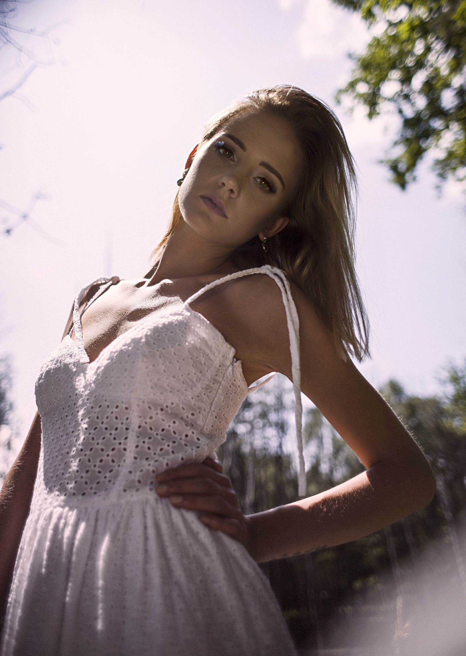 Portret kobiety w białej, ażurowej sukience na tle rozmytej zieleni drzew i jasnego nieba, ujęcie z dołu.