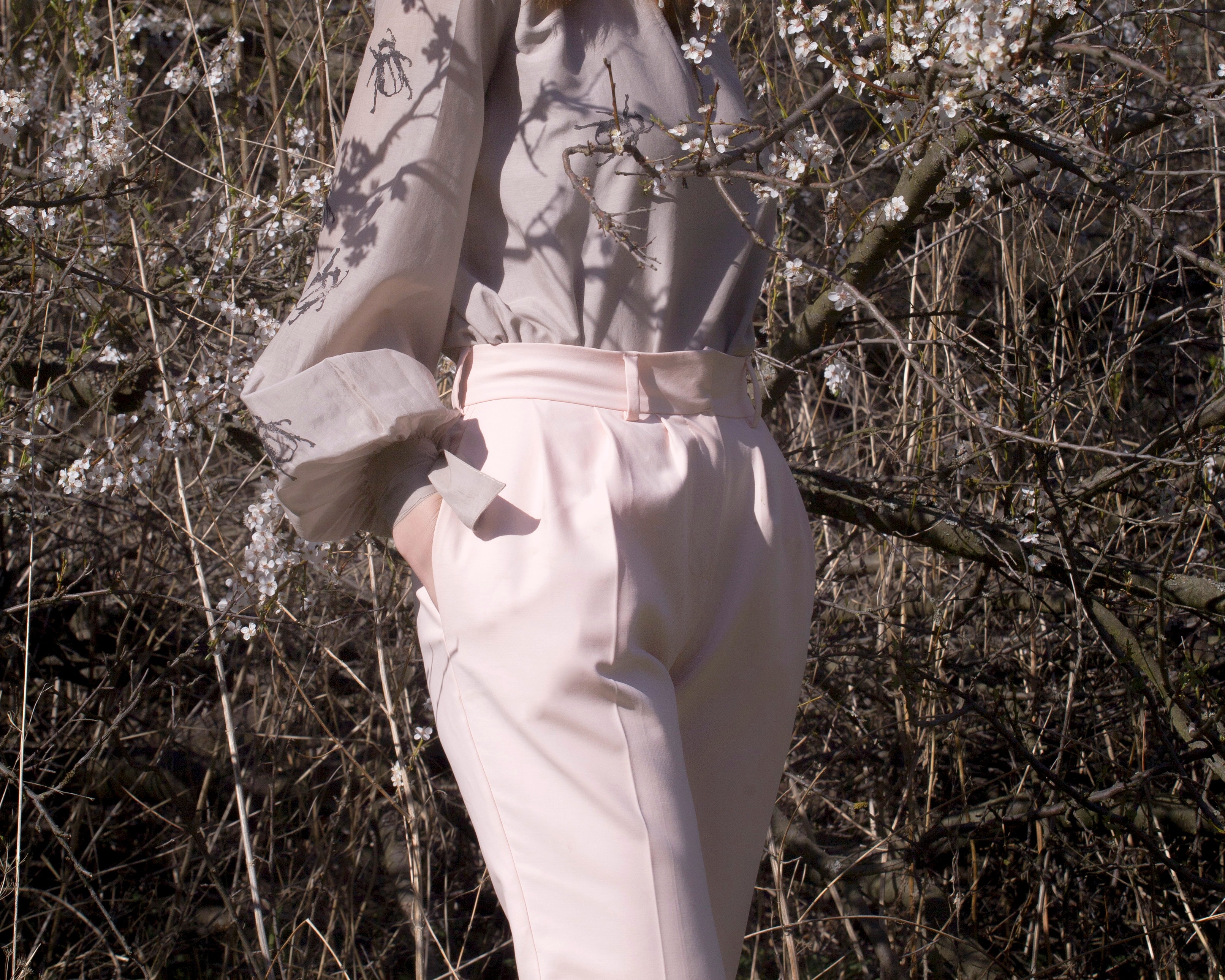 Prezentacja sylwetki w pastelowej bluzce z nadrukiem i różowych spodniach na tle kwitnących gałęzi.