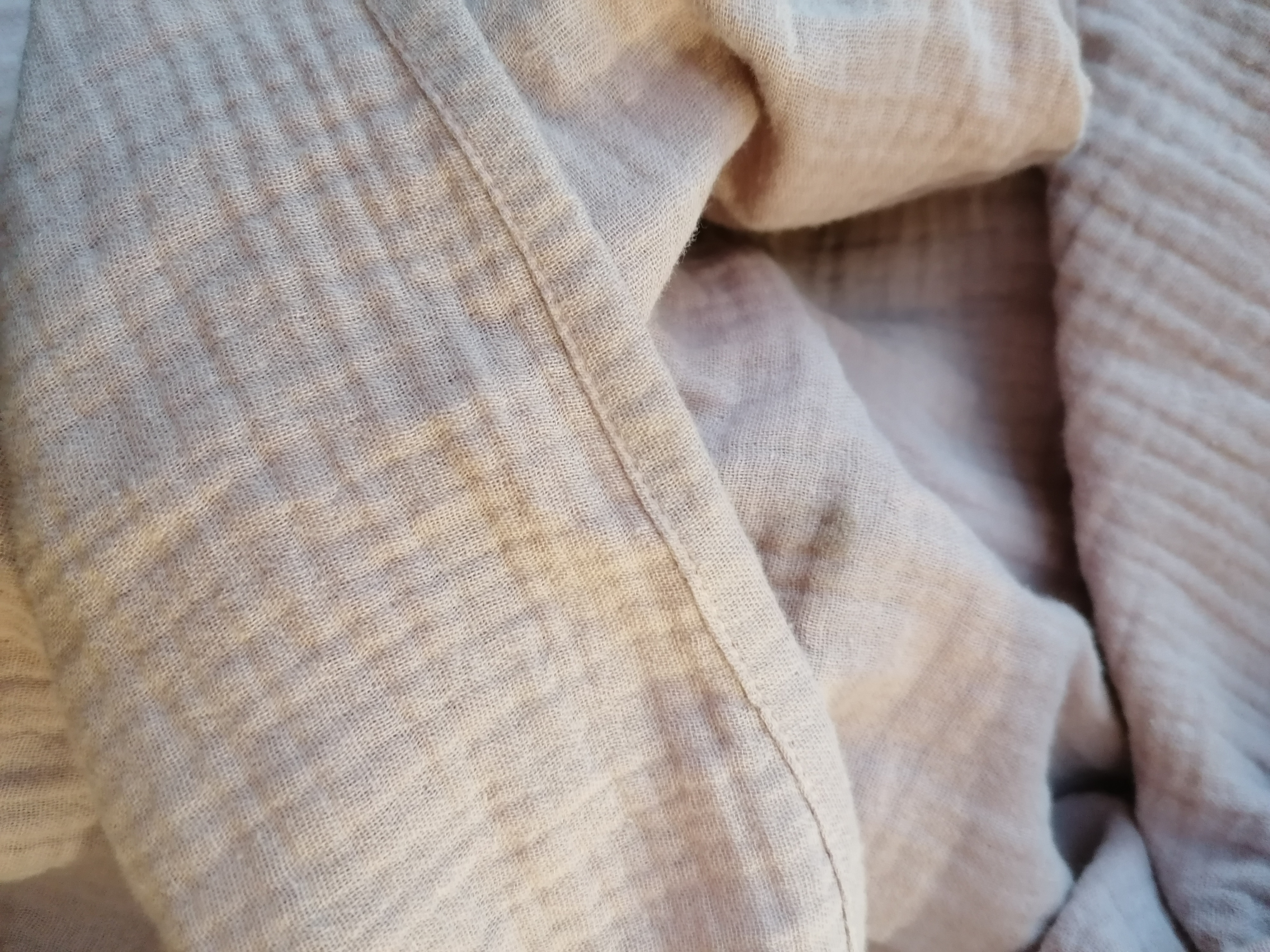 Detale beżowej, muślinowej tkaniny z widocznym szwem, ukazujące jej delikatną fakturę i miękkość.