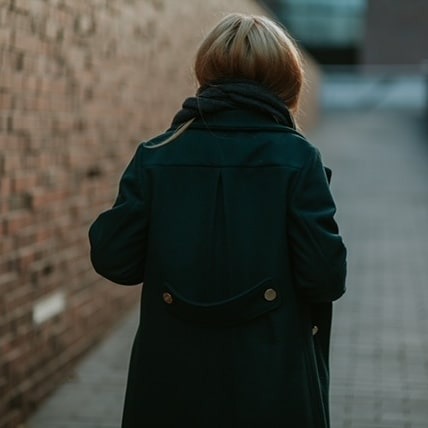 Tył ciemnozielonego płaszcza damskiego z ozdobnymi guzikami, widok z tyłu na blond włosy i szal, na tle ceglanej ściany i chodnika.