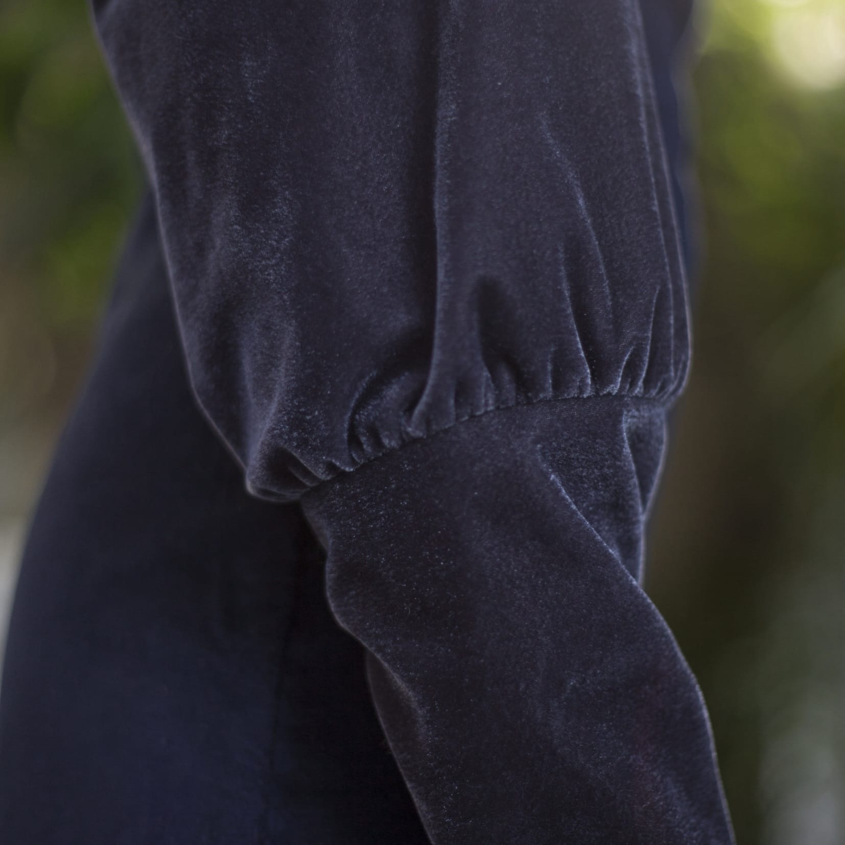 Zbliżenie na rękaw sukienki z ciemnego aksamitu z marszczeniem przy mankiecie, na rozmytym zielonym tle.