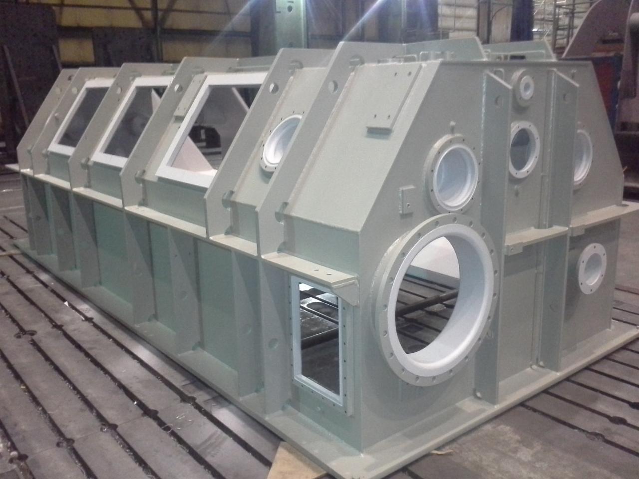 Duża, szara, metalowa konstrukcja z otworami o różnych kształtach (okrągłe i kwadratowe) na metalowej podłodze w hali produkcyjnej. Konstrukcja wydaje się być nowa i świeżo pomalowana.