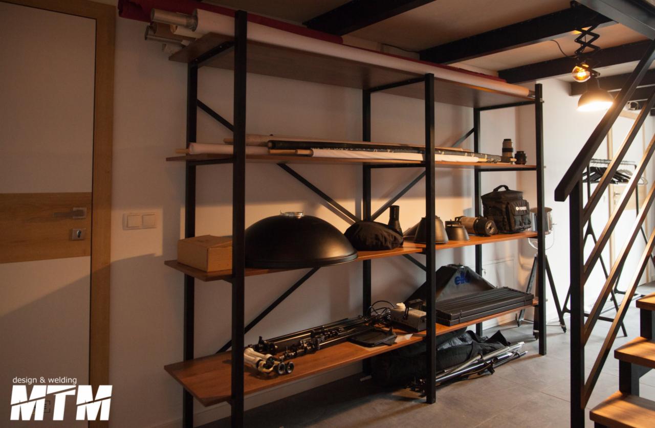 Metalowy regał z drewnianymi półkami, wypełniony sprzętem fotograficznym i oświetleniowym, widoczny w tle fragment schodów i drzwi.