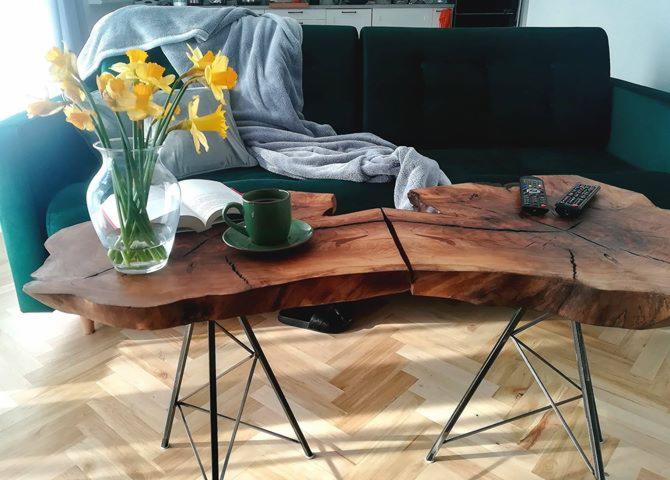 Stolik kawowy z blatem z naturalnego drewna o nieregularnym kształcie, wsparty na metalowych nogach, z wazonem z żółtymi narcyzami, filiżanką kawy i książką na blacie, ustawiony w salonie z zieloną...