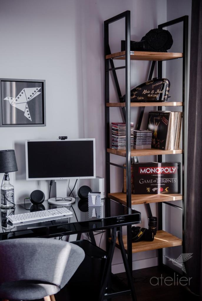 Nowoczesny pokój z biurkiem z czarnym szklanym blatem, białym komputerem i regałem w stylu loft z drewnianymi półkami, na których znajdują się płyty CD, gry planszowe i pamiątki.
