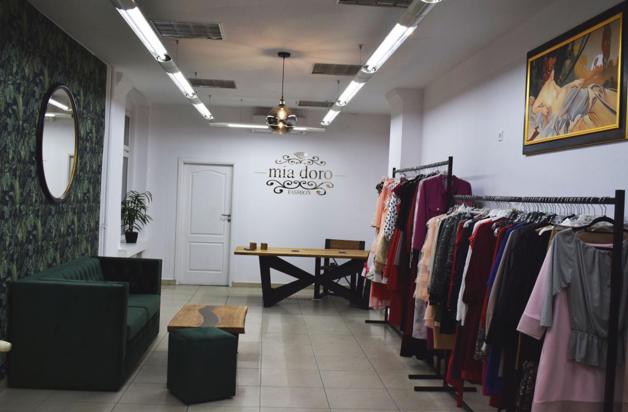 Wnętrze butiku odzieżowego z zieloną sofą, stolikiem kawowym z żywicą, wieszakami z ubraniami i logo marki na ścianie.