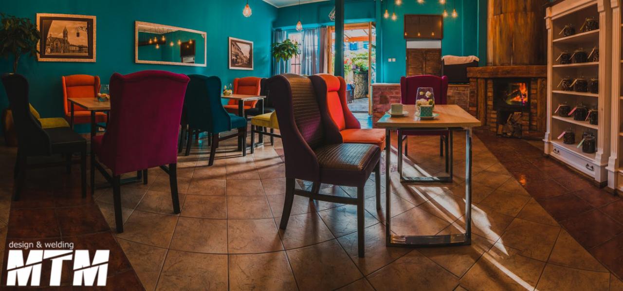 Turkusowe wnętrze kawiarni z kolorowymi krzesłami, drewnianymi stolikami na metalowych nogach, kominkiem i regałem z słoikami, oświetlone wiszącymi lampami.