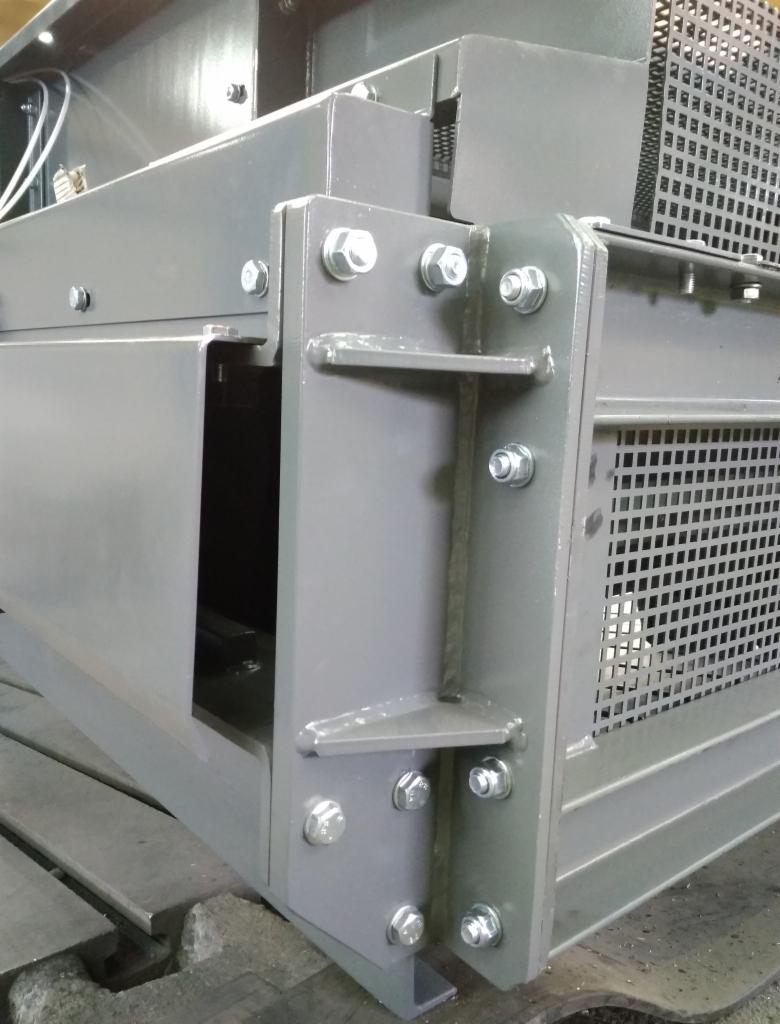 Szary metalowy element konstrukcyjny z perforowaną blachą, połączony śrubami, widoczny z bliska, z fragmentem drewnianej podstawy.