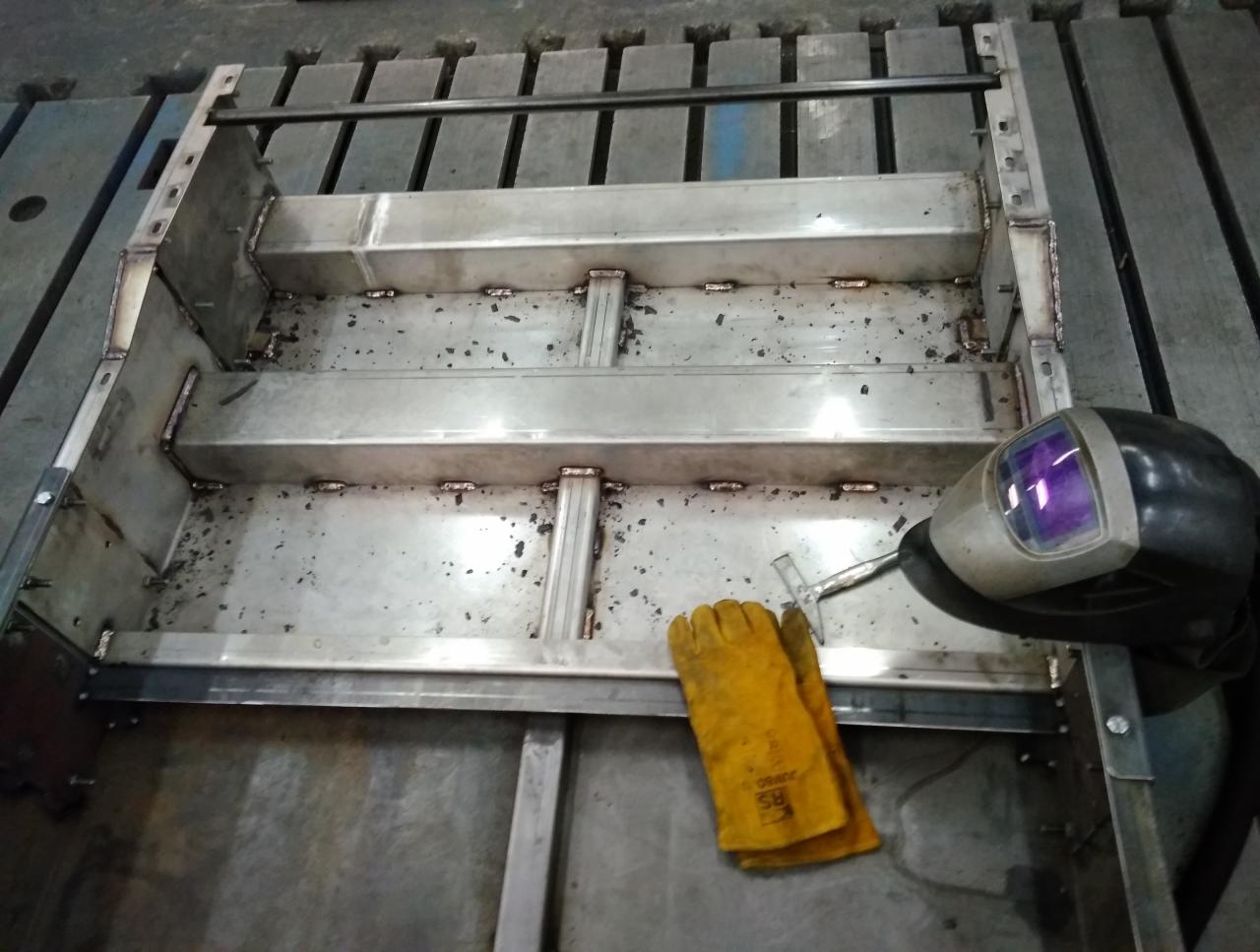 Konstrukcja metalowa ze śladami spawania, żółte rękawice robocze i maska spawalnicza z fioletowym wizjerem leżą na powierzchni roboczej.