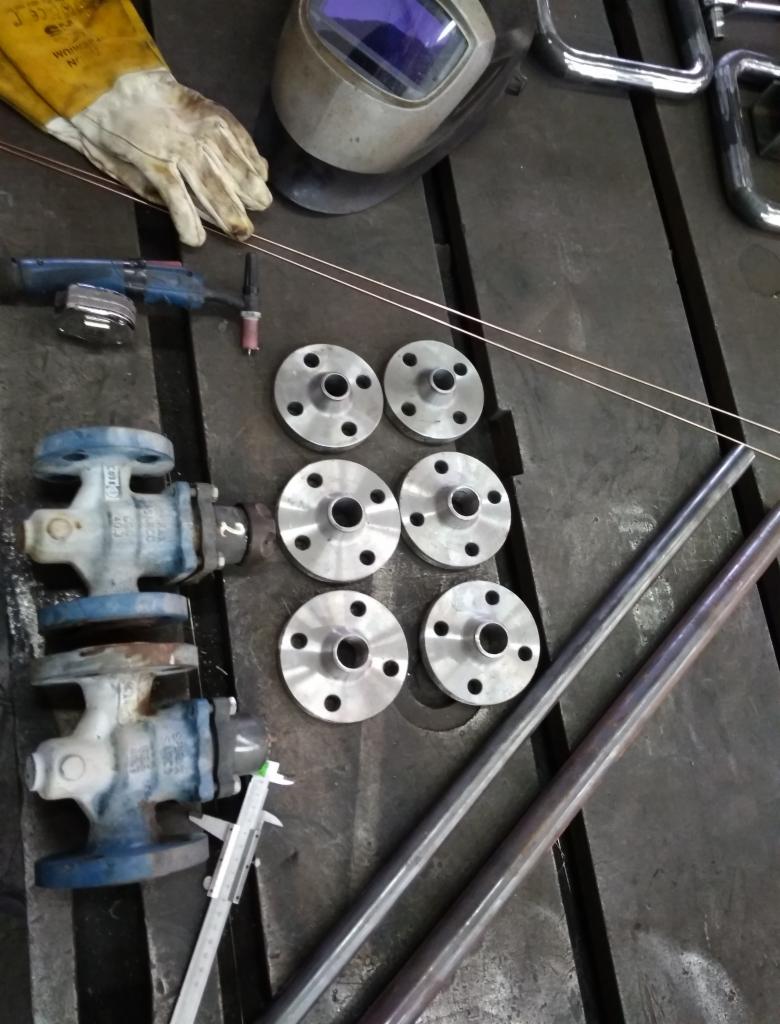 Przygotowanie do spawania: maska spawalnicza, rękawice ochronne, palnik, rury, zawory i metalowe kołnierze ułożone na warsztatowym stole. Widoczny suwmiarka.