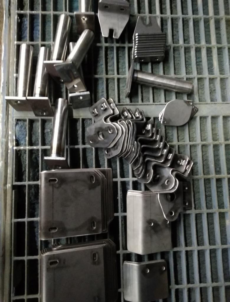 Różnorodne metalowe elementy i detale, ułożone na metalowej kratce, widoczne ślady obróbki.