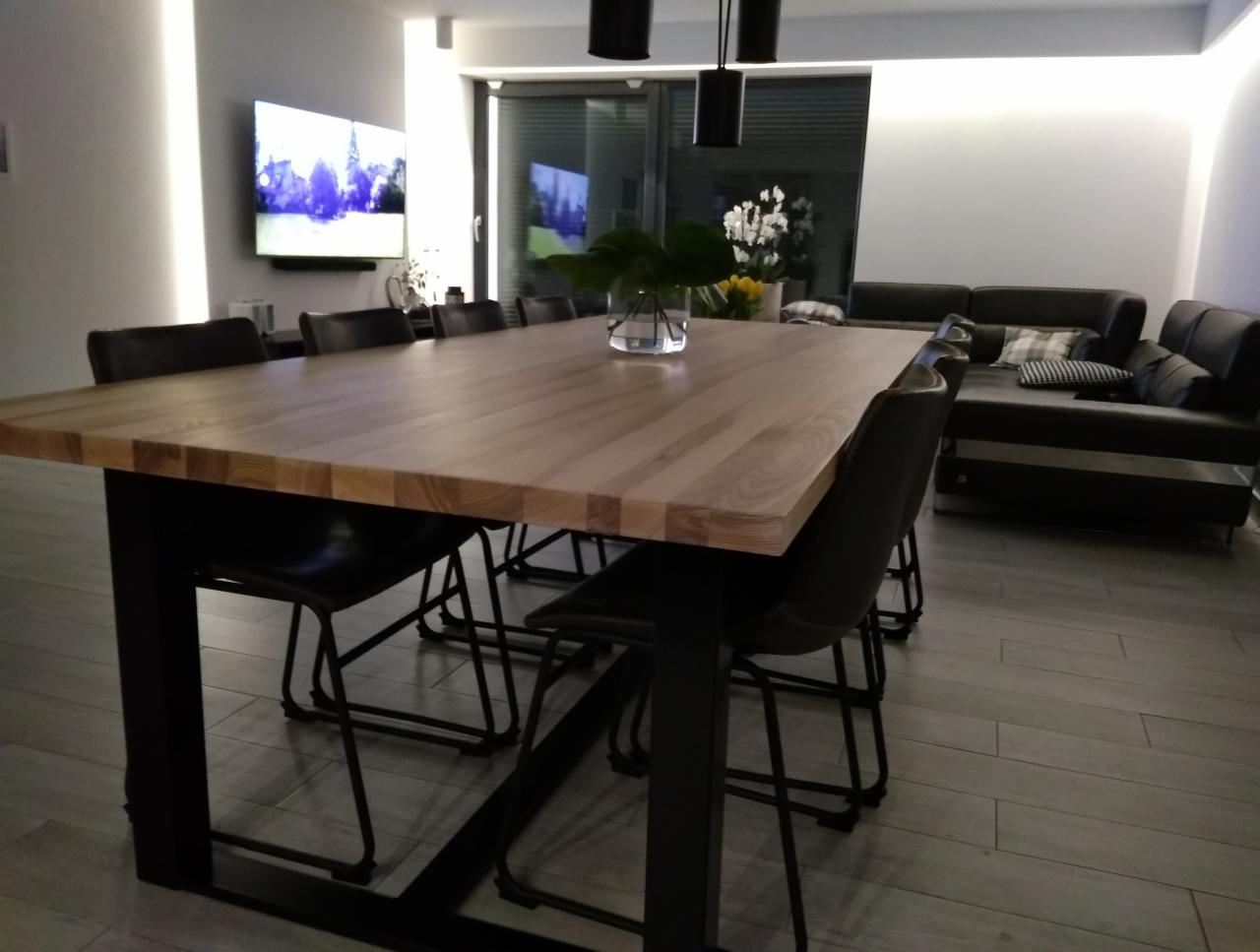 Nowoczesna jadalnia z dużym, jasnym stołem z litego drewna na metalowych nogach, otoczonym czarnymi krzesłami, w tle minimalistyczny salon z szarą sofą i telewizorem.