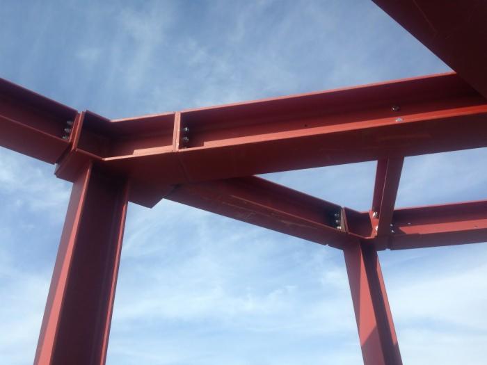Konstrukcja stalowa budynku w trakcie montażu, czerwone elementy połączone śrubami na tle błękitnego nieba z chmurami.