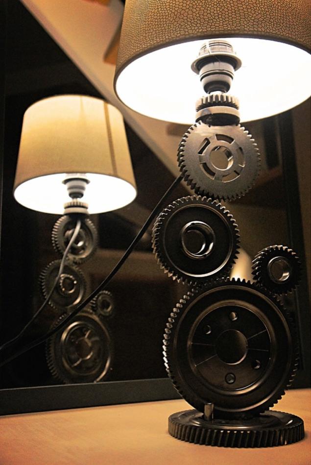 Lampa stołowa w stylu industrialnym, wykonana z metalowych kół zębatych, z beżowym abażurem, odbijająca się w lustrze.