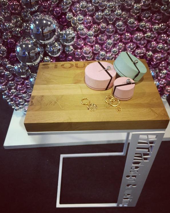 Drewniany stolik z metalowymi nogami prezentuje okrągłe pudełka w pastelowych kolorach oraz złotą biżuterię na tle ściany z dekoracyjnych, srebrnych kul.