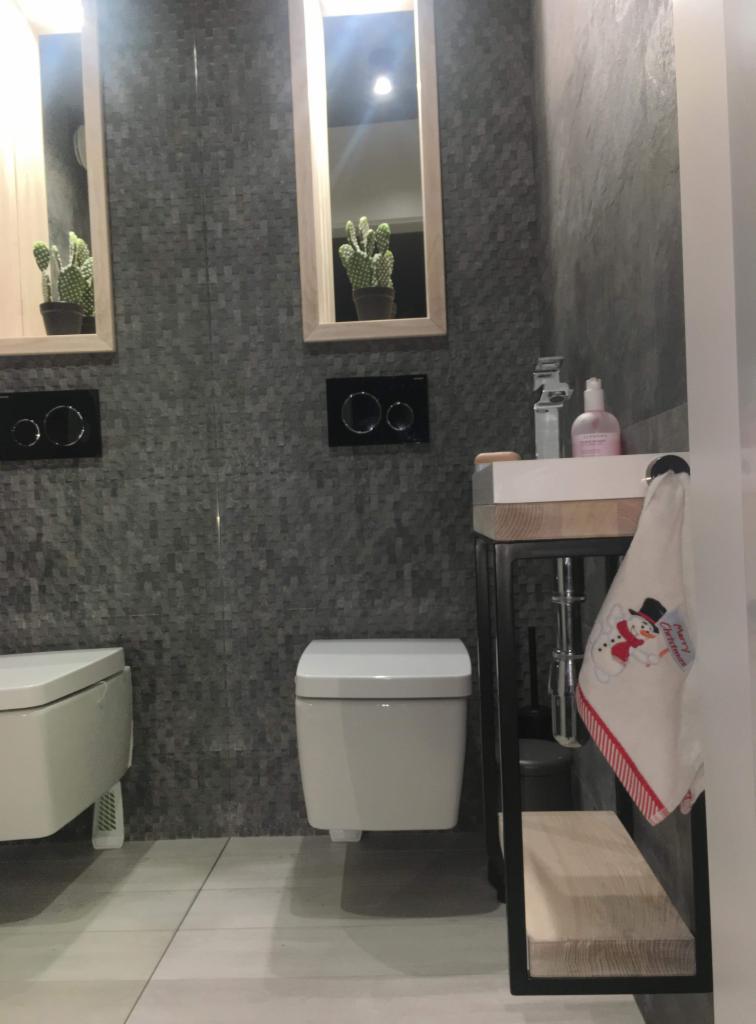 Nowoczesna łazienka z szarą mozaikową ścianą, wiszącą toaletą, umywalką na metalowym stelażu z drewnianymi półkami, lustrami z kaktusami i ręcznikiem z motywem świątecznym.