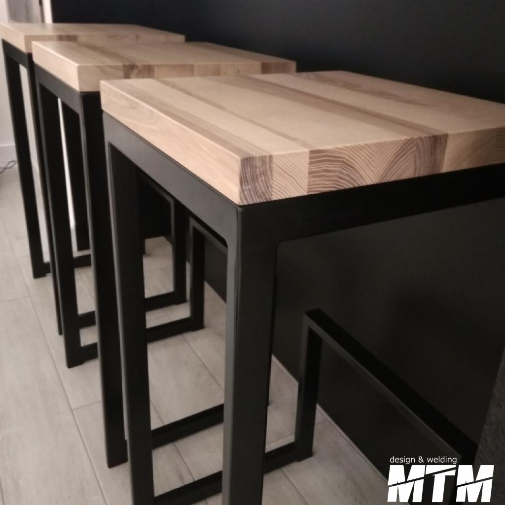 Trzy identyczne hokery z drewnianymi blatami o wyraźnym usłojeniu i czarnymi metalowymi nogami o kwadratowym przekroju, ustawione jeden za drugim na jasnej podłodze.