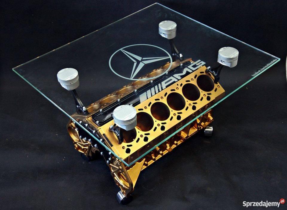 Stolik kawowy wykonany z silnika samochodowego AMG, z szybą z wygrawerowanym logo Mercedesa, wsparty na tłokach, złoty blok silnika na czarnym tle.