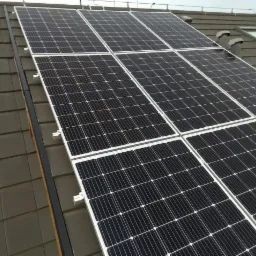 Czarno-białe panele słoneczne zamontowane na brązowym dachu z dachówki ceramicznej, widoczne wsporniki mocujące i fragment okna dachowego.