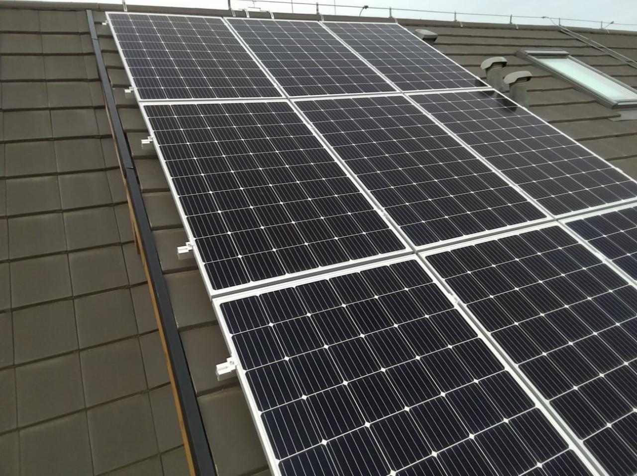 Czarno-białe panele słoneczne zamontowane na brązowym dachu z dachówki ceramicznej, widoczne wsporniki mocujące i fragment okna dachowego.