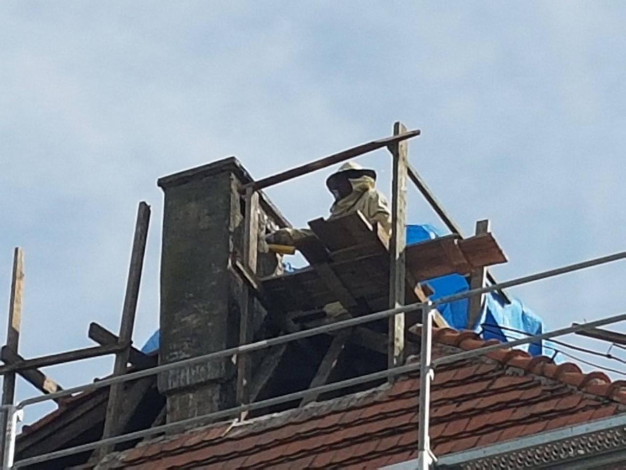 Osoba w kombinezonie ochronnym pszczelarza usuwa gniazdo os z komina na dachu domu, widoczne rusztowanie i plandeka zabezpieczająca.
