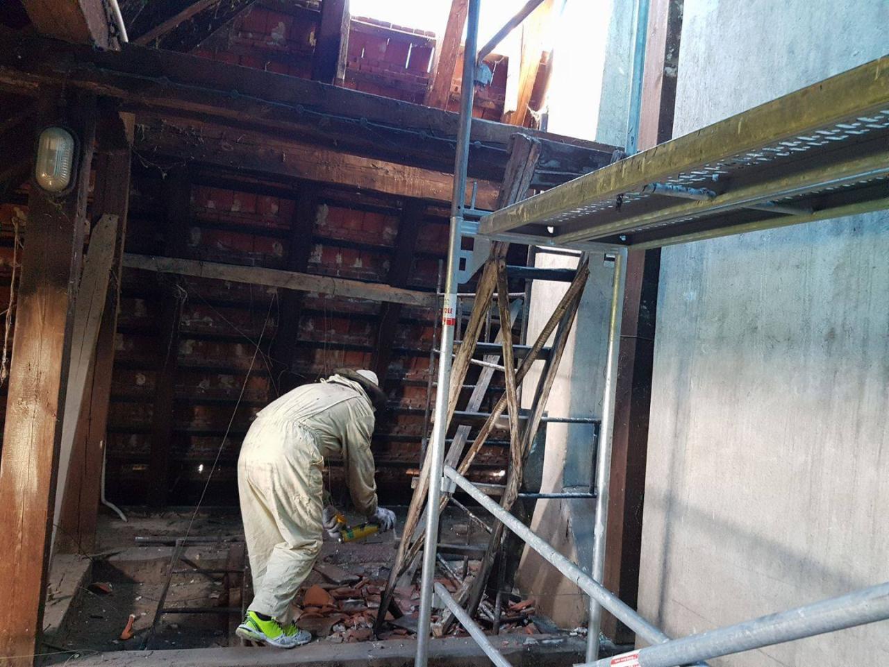 Pracownik w kombinezonie ochronnym i jaskrawych butach aplikuje preparat na drewnianą konstrukcję poddasza z rusztowania.