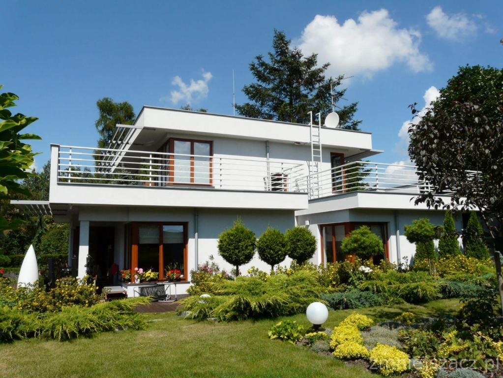 Nowoczesny, dwukondygnacyjny dom z białymi ścianami, płaskim dachem i balkonem z metalową balustradą, otoczony zadbanym ogrodem z kulistymi krzewami i kwitnącymi rabatami.